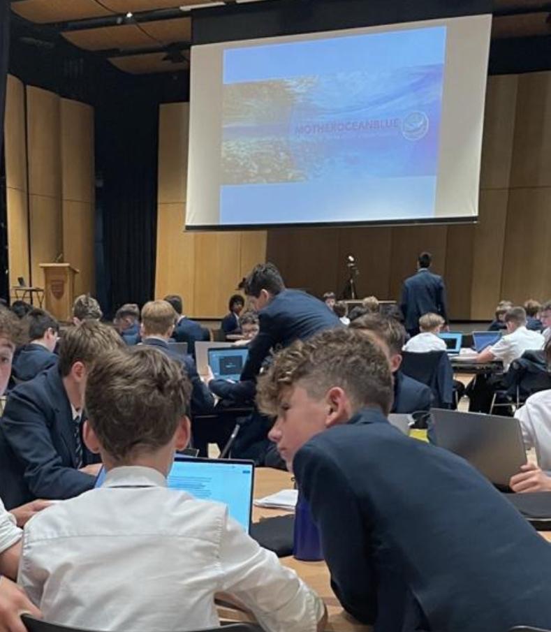 Students working on laptops during an Ocean Academy session in a school hall, the MotherOceanBlue title slide on screen